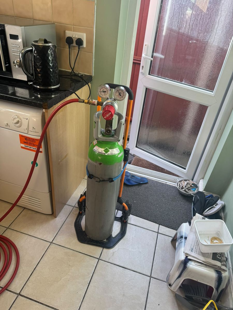 Tracer gas cylinder with regulator on a trolley in a kitchen, red hose running across the floor
