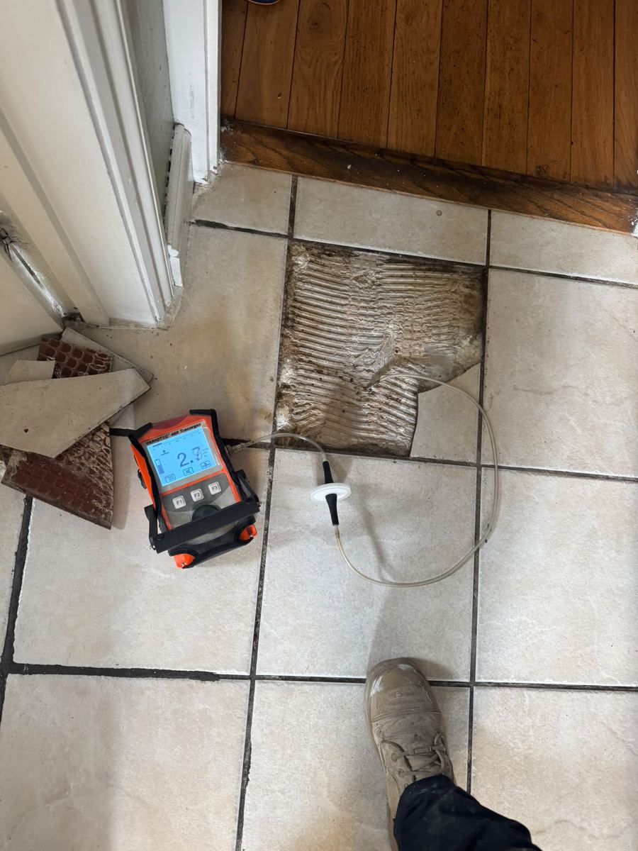 Tracer gas detector reading beside a lifted floor tile, pinpointing the leak